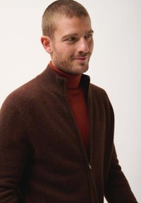 Cardigan marrone con zip realizzato in lana texturizzata, con colletto alto, indossato sopra un dolcevita ruggine. Presenta una vestibilità aderente e un tessuto morbido.