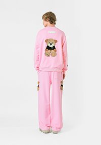 Conjunto de sudadera y pantalones de forro polar rosa que presenta un diseño de oso de peluche con una camiseta negra, aplique texturizado en la parte trasera y los lados.