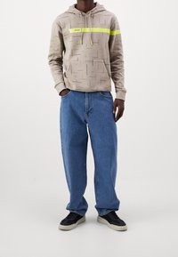 Ljusbeige hoodie med geometriskt mönster och en lysande gul rand, kombinerad med lösa blå denimjeans och svarta sneakers.