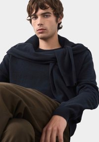 Maglione lavorato a maglia blu navy con collo drappeggiato, abbinato a pantaloni marrone chiaro. Tessuto morbido, vestibilità rilassata e dettagli di design minimi.