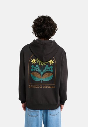 Personne aux cheveux bouclés portant un sweat à capuche noir avec un motif végétal coloré et le texte "Balance of Opposites" dans le dos.