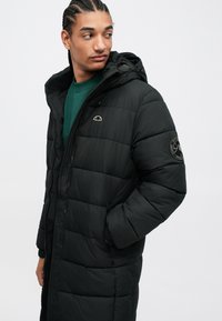 Άνδρας με πλεξούδες, φορώντας μαύρο καπιτονέ μπουφάν με κουκούλα πάνω από έναν πράσινο σκούφο, κοιτάζει στο πλάι με το ένα χέρι στην τσέπη του μπουφάν.