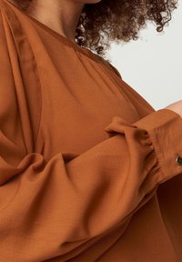 Blusa color ruggine caratterizzata da un tessuto morbido e testurizzato, design oversize, maniche ampie con polsini arricciati e delicate pieghe sul collo.