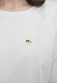 White sweatshirt featuring a small embroidered design of two figures on a cloud, with a soft texture and simple raglan sleeves.
