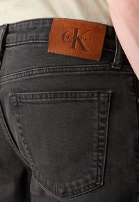 Primo piano della tasca posteriore di jeans denim grigio scuro con una patch in pelle marrone del marchio Calvin Klein sulla vita.