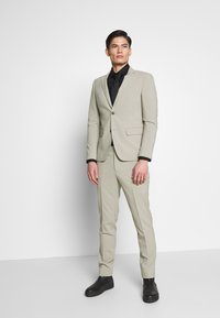 Completo grigio chiaro con vestibilità su misura, blazer monopetto, camicia nera sotto e scarpe nere. Tessuto liscio, dettagli minimi.