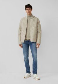 Mann in einer beige gesteppte Jacke über einem beigen Rollkragenpullover, blauen Jeans und weißen Sneakern, der vor einem schlichten grauen Hintergrund steht.