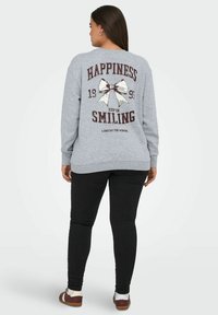 Felpa grigia con un design stampato che presenta le parole "FELICITÀ," un'illustrazione di un fiocco e "CONTINUA A SORRIDERE." Abbinata a leggings neri.