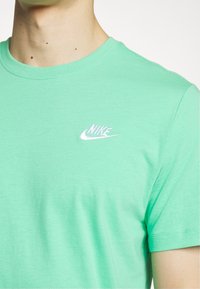 Detailní záběr na osobu oblečenou v světle zeleném tričku Nike s bílým vyšívaným logem na hrudi, zobrazuje krk a horní část trupu.