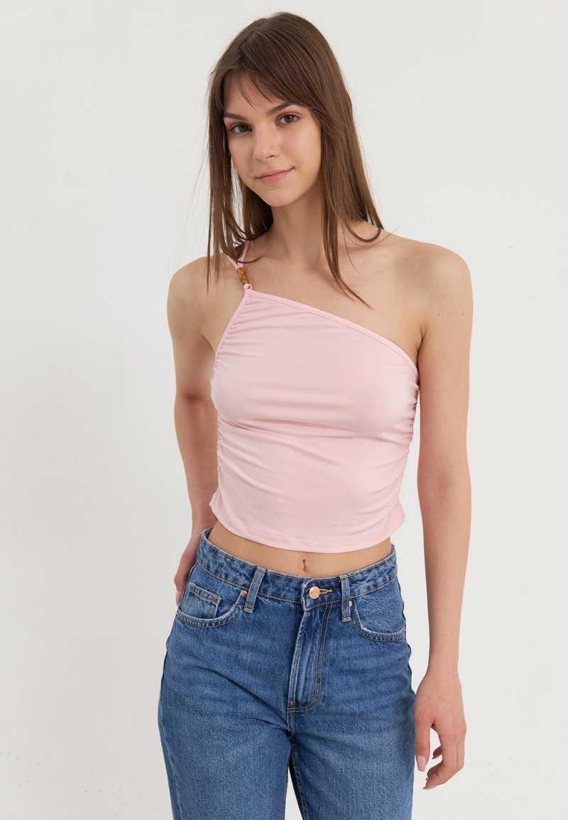 Top sans manches rose clair avec un design asymétrique, texture plissée sur le côté et un bouton décoratif à l'épaule. Associé à un jean bleu.