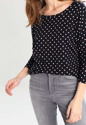 Top a pois blu navy realizzato in tessuto leggero, con scollatura ampia e maniche a tre quarti, abbinato a jeans grigi aderenti.