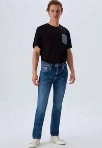 Jovem de pé, vestindo uma t-shirt preta com um bolso cinza, jeans azuis e ténis brancos, contra um fundo claro liso.
