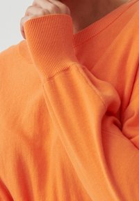 Pomarańczowy sweter z dekoltem w serek, ściągaczami przy mankietach i gładką fakturą, wykonany z miękkiej dzianiny o luźnym kroju.