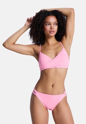 Kobieta o kręconych włosach w jasnoróżowym bikini, pozująca z uniesionymi ramionami, patrząca lekko na bok na jednolitym białym tle.