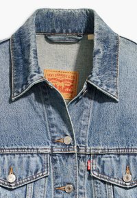 Giacca in denim azzurro chiaro con colletto, doppie tasche sul petto, bottoni argentati, etichetta beige con testo rosso. Tessuto testurizzato e cuciture a contrasto.