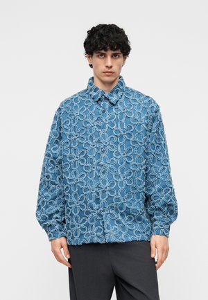 Junger Mann trägt ein blaues Hemd mit Knöpfen, das ein erhabenes Blumenmuster in Form von Schlaufen aufweist, und dunkle Hosen vor einem schlichten weißen Hintergrund.