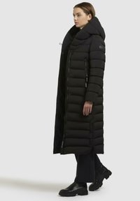 Lungo cappotto imbottito nero con colletto alto, cuciture orizzontali e tasche laterali con zip. Indossato sopra pantaloni scuri e stivali.