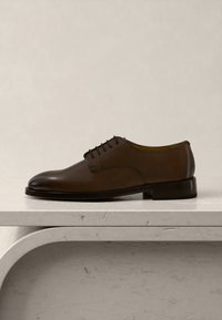 Chaussure de ville à lacets en cuir brun foncé unique, présentée sur une surface en marbre clair avec un fond uni.