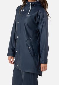 Femme portant un imperméable bleu marine à capuche avec boutons-pression, poches à rabat et cordons blancs, debout sur un fond blanc.