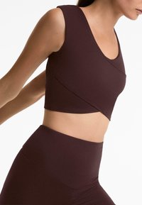 Tamno smeđi atletski crop top s izrezom u obliku uvale, bez rukava, s dijagonalnim detaljima šava, u kombinaciji s tajicama viskog struka.