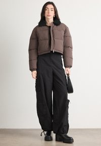Brązowa krótka kurtka puchowa z czarnym futrzanym kołnierzem, w połączeniu z czarnymi szerokimi spodniami cargo. Modelka trzyma małą czarną torebkę.