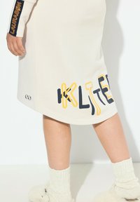 Crème kleurig kledingstuk met opvallende tekst in geel en marineblauw. Bevat een logo en geribde manchetten. Zachte textuur met een relaxte pasvorm.
