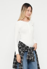Biely dlhý rukáv henley top s gombíkovou légou, v kombinácii s viazanou čiernou a šedou kockovanou košeľou v páse a modrými džínsami.