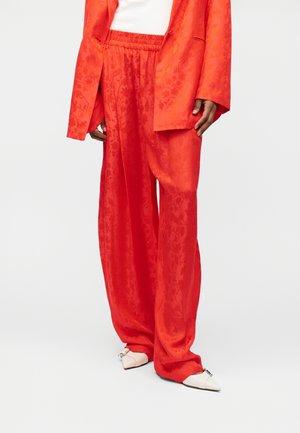 Oseba, ki nosi široke, svetlo rdeče hlače z rožnatim vzorcem in ustrezno srajco, kombinirano z belimi ostrimi čevlji s srebrnim zaponkom.