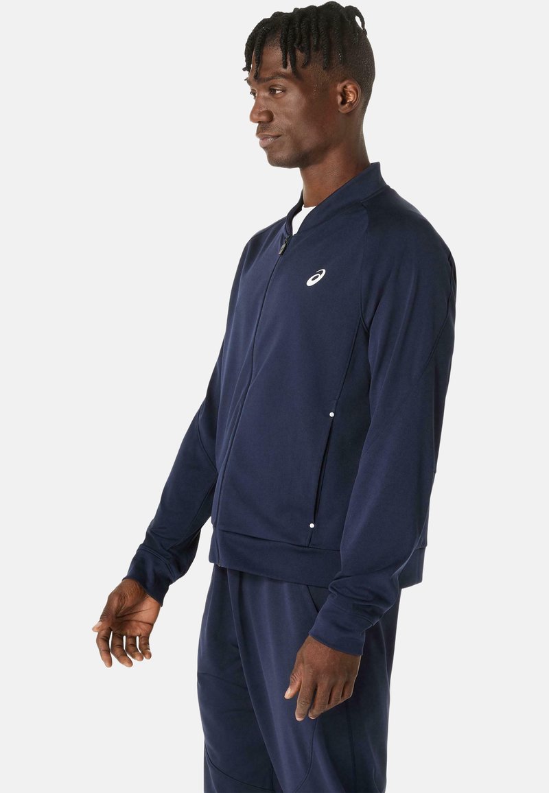 Giacca sportiva blu navy in tessuto liscio, con zip frontale, colletto rialzato e logo discreto sul petto. Maniche lunghe e design aderente.