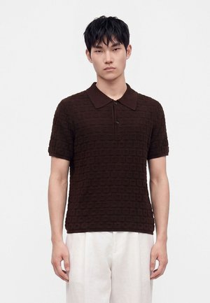 Jeune homme aux cheveux courts noirs porte un polo marron foncé texturé à manches courtes et un pantalon blanc ample sur un fond clair uni.