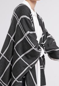 Chal gris carbón con un patrón de cuadrícula blanca, hecho de una tela suave y texturizada. Colocado elegantemente sobre una camisa blanca, mostrando su forma oversized.