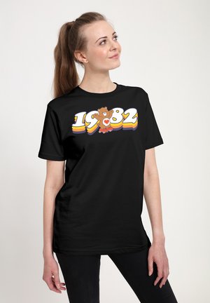 Sort T-shirt med et farverigt grafik af en tegneserie-bjørn og året "1982" i fed, legesyg skrifttype. Blødt stof med en afslappet pasform.