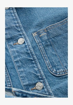 Jasnoniebieska kurtka dżinsowa z srebrnymi guzikami, podwójnymi szwami i kieszenią na piersi z klapką. Materiał jest miękki i zużyty.