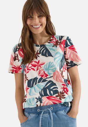 Femme souriante aux cheveux bruns portant une chemise à manches courtes à imprimé floral tropical et un pantalon en jean bleu à cordon, les mains dans les poches.
