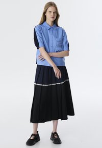 Camicetta blu a maniche corte con taschino e dettagli neri, abbinata a una gonna midi plissettata blu navy e nera con bordo in pizzo. Sneakers nere.