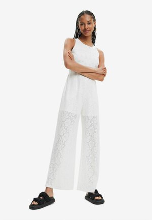 Jumpsuit bianco in pizzo con gambali larghi, caratterizzato da un design senza maniche e intricati motivi floreali. Indossato con sandali neri.