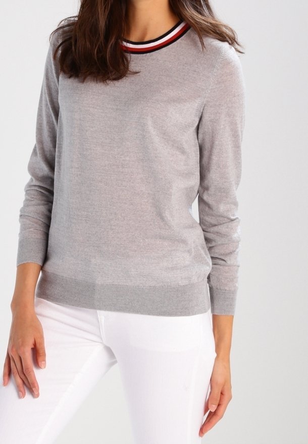 Femme portant un pull à manches longues gris clair avec un col rayé rouge, blanc et noir et un pantalon blanc, debout devant un fond uni.