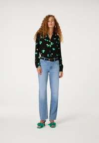 Schwarzes Hemd mit knöpfen und grünem Blätterdruck, blaue hochgeschnittene Jeans und grüne Sandalen mit Schleife. Lockige Haare und trägt Creolen.