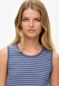 Jonge vrouw met lang, blond haar, in een mouwloos blauw en paarse gestreepte top, die recht vooruit kijkt met een neutrale uitdrukking.