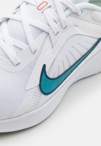 Primer plano de una malla de zapatilla deportiva blanca con el swoosh en teal de Nike y cordones blancos atados en un patrón entrelazado.
