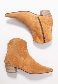 Botines de ante en tono beige con puntera afilada y cremallera lateral. Presentan un tacón de madera apilada y detalles de costura en la parte superior.