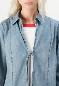 Svetlo modra denimska srajca s ovratnikom, odprta spredaj z zankico, ter vidnimi detajli šivanja, v slojih nad belim rebrastim vrhom z rdečimi poudarki.