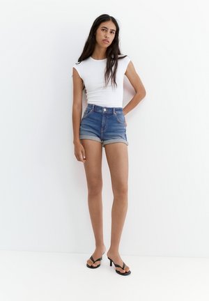 Femme aux longs cheveux foncés portant un haut blanc sans manches, un short en jean retroussé et des sandales noires à talons, debout contre un mur blanc.
