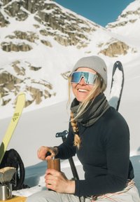 Lächelnde Frau in Winterkleidung und Sonnenbrille mahlt Kaffee im Freien in einer verschneiten Berglandschaft mit Skiern und Ausrüstung in der Nähe.