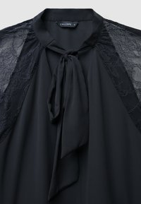 Blusa nera con dettagli in pizzo sulle spalle, un collo con lacci e una texture liscia e morbida. L'etichetta del marchio reca la scritta "CALLIOPE."