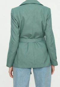 Green belted blazer with a notched collar, structured shoulders, and a smooth texture. The back features a pleated design and is tailored fit.
