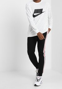 Persoon gekleed in een witte blouse met lange mouwen en stippen, met een zwart logo, zwarte broek met rode en witte zijkanten, en witte sneakers met zwarte accenten.