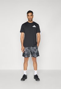 T-shirt noir à col ras du cou avec un graphique de montagne sur la poitrine, associé à un short gris tie-dye. Le modèle porte des baskets noires et des chaussettes blanches.