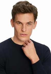 Marineblaue Rundhals-Strickjacke mit gerippten Kanten, getragen von einer Person mit kurzen, texturierten braunen Haaren, die Hand am Kinn, neutraler Hintergrund.