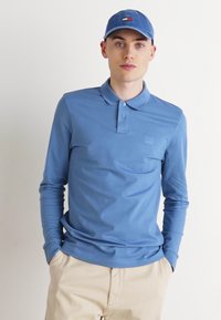 Ilgų rankovių šviesiai mėlynos spalvos polo marškiniai su apykakle ir trimis mygtukais, derinami su atitinkančia skrybėle su mažu logotipu. Modelis dėvi smėlio spalvos kelnes.
